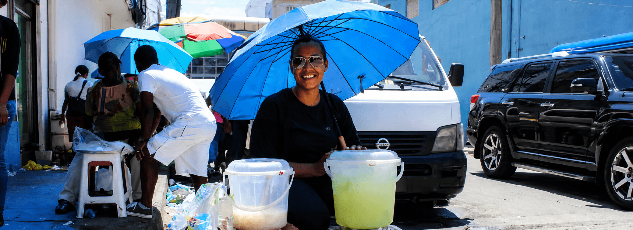 Vendedora ambulante bajo una sombrilla azul en una calle urbana, imagen usada para representar actualidad y movilidad humana